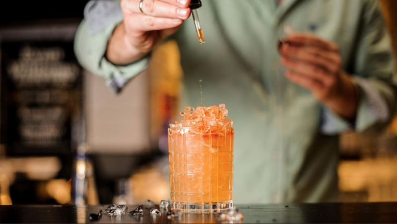 A hand drips some bitters from a dropper into a cocktail. The cocktail is peach-hued and has lots of crushed ice. 