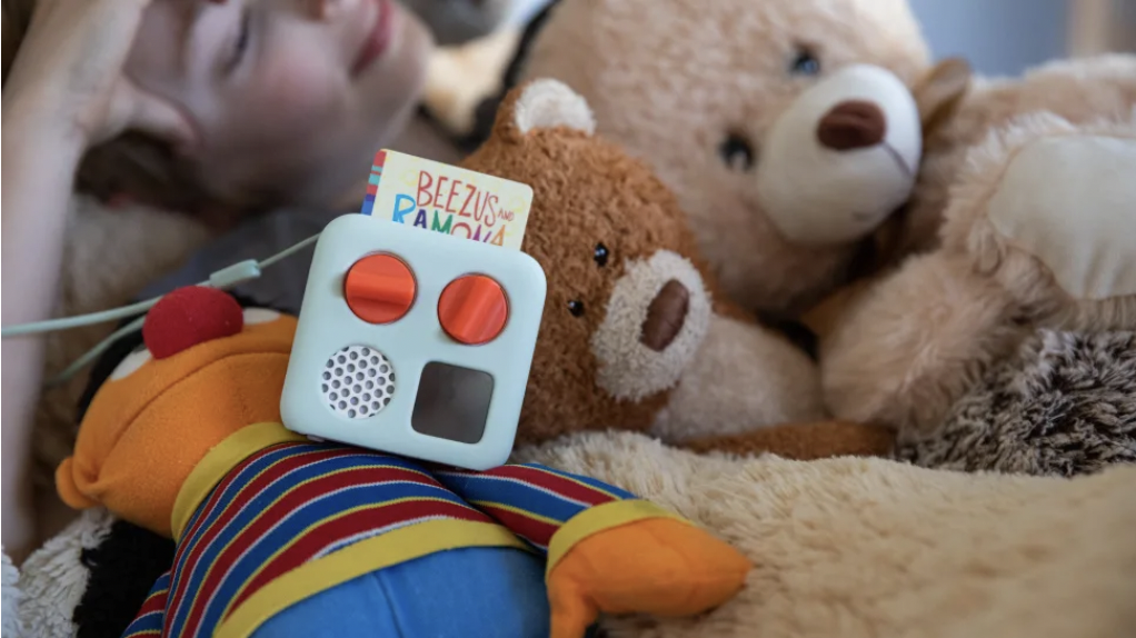 A child lies down on a bed of stuffed animals with a Yoto Mini kids' audio player.