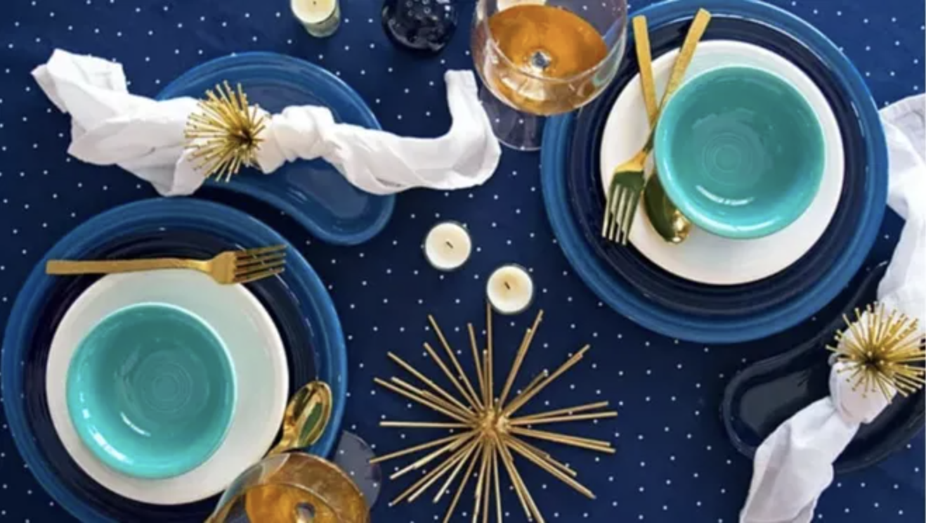 A deep blue background with settings of Fiestaware in blue, teal, and white. There are accents of gold, including gold forks and spoons.