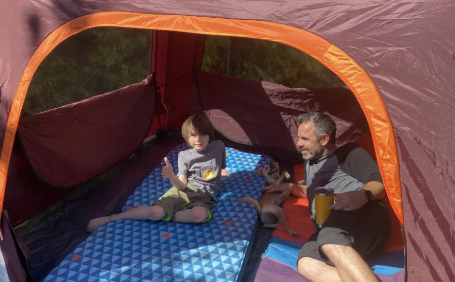 A child gives a "thumbs up" in a tent while his dad and puppy look on.