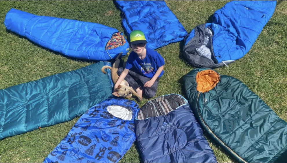 A child sits in the center of blue and green sleeping bags fanned out like a star. He is sitting on grass with his puppy at his feet.
